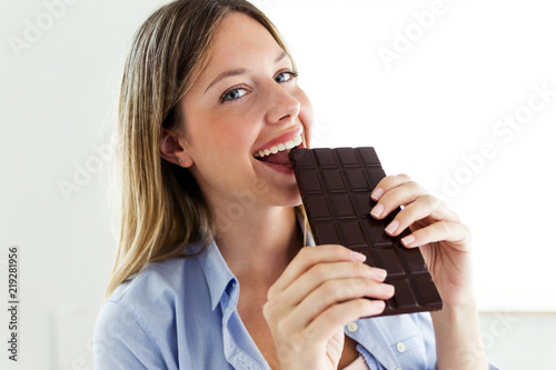 Beautiful young woman biting chocolate bar at home.