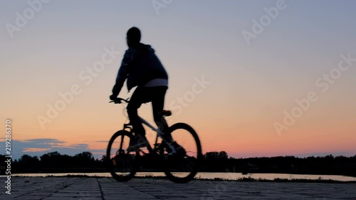 A teenager on a two-wheel self-balancing hoverboard and a guy on a bicycle ride past each other near the pond along the embankment at dawn. Sports lifestyle. Silhouette of a boys in the morning light.