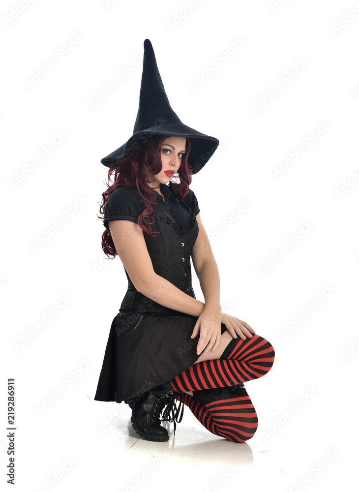 full length portrait of red haired girl wearing black witch costume and pointy hat. seated pose, isolated on white studio background. full length portrait of red haired girl wearing black witch costume and pointy hat. seated pose, isolated on white studio background.