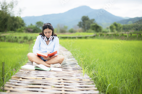 Asian young woman enjoy readbooks on wooden bridge in nature rice farm; her is feeling relax and easy listening music online from mobile device in the meanwhile; Relax and happy concept. photo