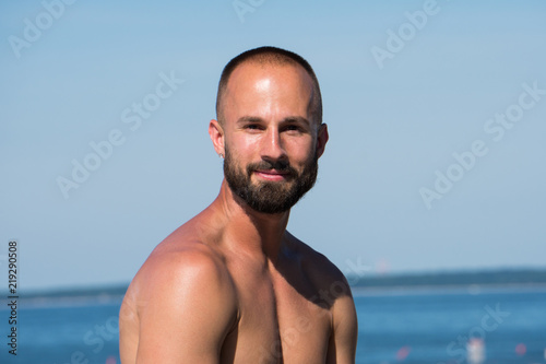 Young handsome athletic adult man with short hair and beard looking into camera while sitting on beautiful tropical beach during hot summer time day. Ocean background scenic view.