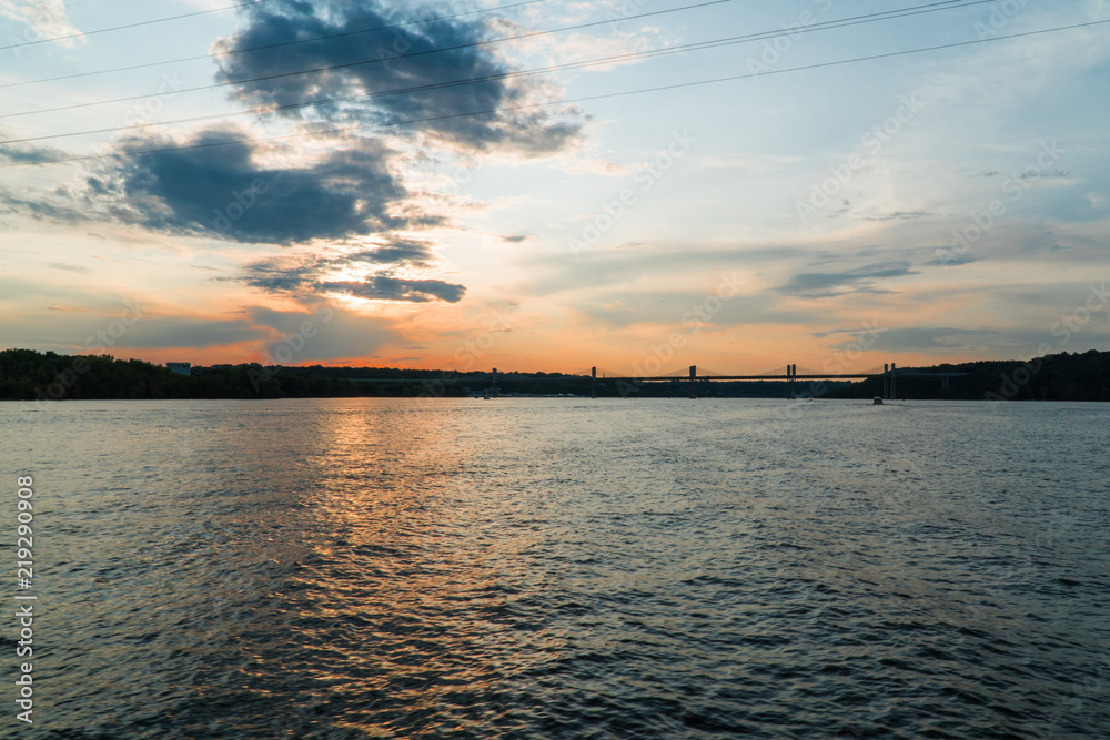Beautiful majestic view of sun setting over a tranquil river partly cloudy summer evening. River coastline on sides connected by bridge in background silhouette