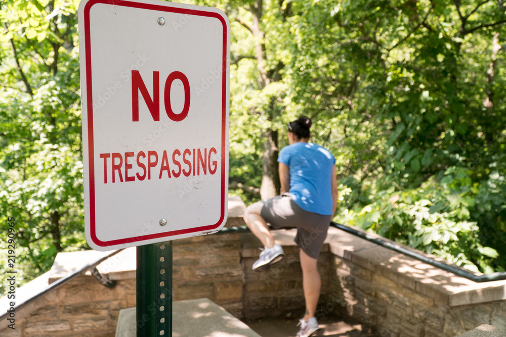 Young woman attempt to jump over railing into restricted area labelled by no trespassing sign. Private property deep woods and forest area illegal to walk past signage
