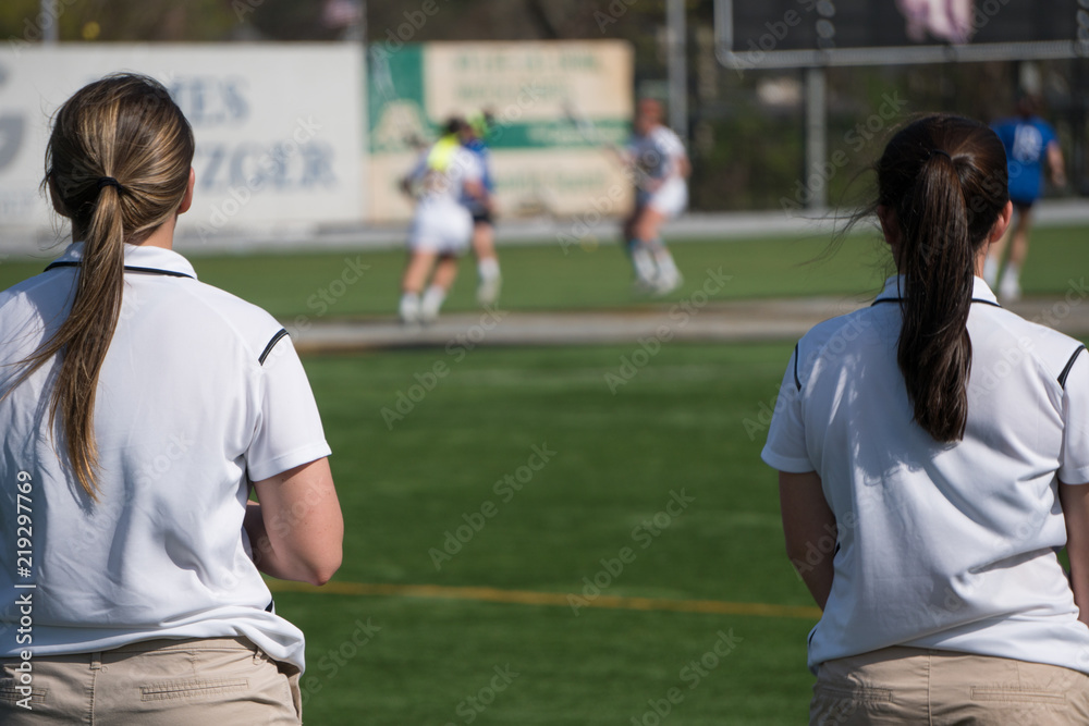 Overlooking a girls lacrosse team sport game two female coaches in uniform colors stand on sideline watching play calling instructions to players on field.