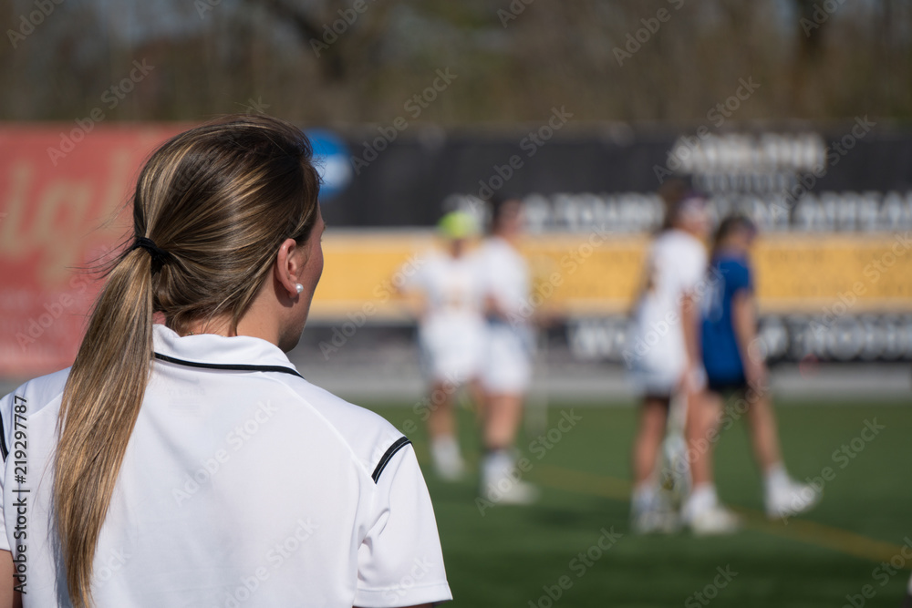 Over the shoulder female head coach on sidelines yelling play call to girl soccer players on field competing tournament match game on bright summer day in outside stadium