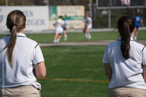 Overlooking a girls lacrosse team sport game two female coaches in uniform colors stand on sideline watching play calling instructions to players on field.