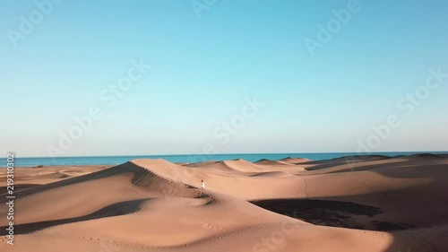 Female in a dress is running over the top of a sand dune in a desert. Beautiful horizontal landscape of desert with blue ocean behind. Perfect vacation. Gran Canaria