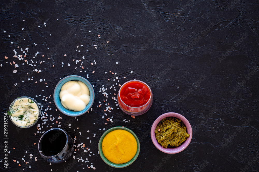 Set of sauces. Ketchup, mayonnaise, mustard, soy sauce, barbecue sauce, pesto, mustard, sour with greenery on black background top view copy space Set of sauces. Ketchup, mayonnaise, mustard, soy sauce, barbecue sauce, pesto, mustard, sour with greenery on black background top view copy space