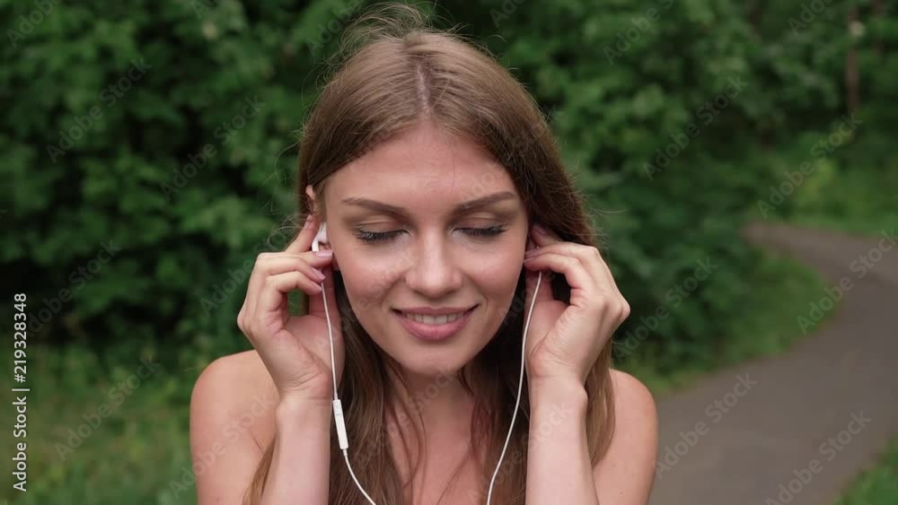 Gorgeous young woman with long dark hair and big eyes plugging in her headphones and listening to music in summer outdoors. Tilt up slow motion medium shot Gorgeous young woman with long dark hair and big eyes plugging in her headphones and listening to music in summer outdoors. Tilt up slow motion medium shot