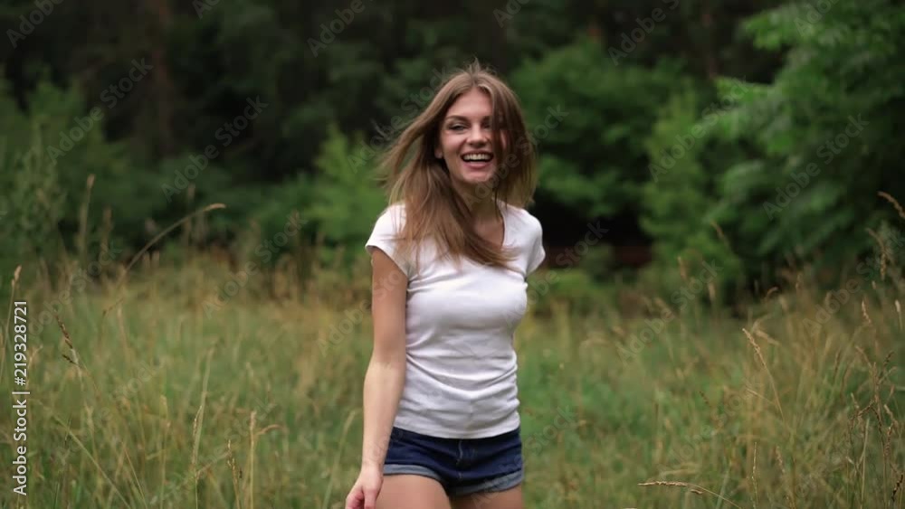 Attractive young woman with long fair hair wearing short jeans shorts and a white t shirt is walking in a summer park and touching the grass. Rear view. Tracking slow motion medium shot Attractive young woman with long fair hair wearing short jeans shorts and a white t shirt is walking in a summer park and touching the grass. Rear view. Tracking slow motion medium shot