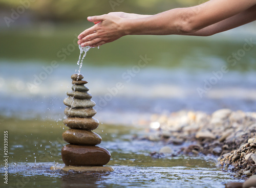 Photography Close-up abstract image of woman hand pouring water on rough natural brown uneven different sizes and form stones balanced like pyramid pile landmark on blurred blue-green copy space background