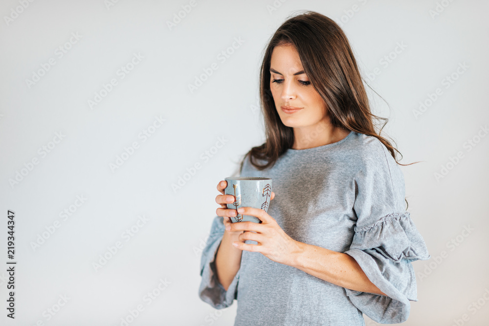 Beautiful woman holding cup of coffee or tea, wearing grey dress, posing against white background Beautiful woman holding cup of coffee or tea, wearing grey dress, posing against white background