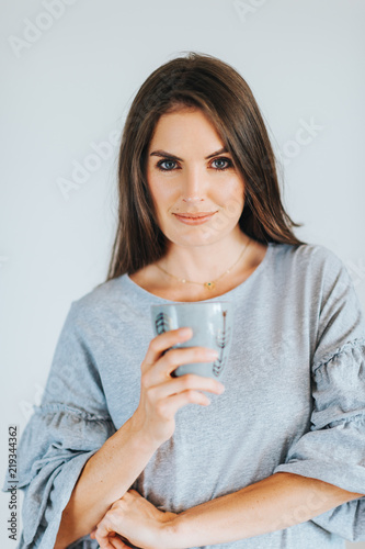Beautiful woman holding cup of coffee or tea, wearing grey dress, posing against white background
