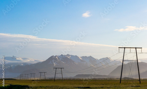 Power poles in an Icelandic landscape at sunset, with snow-capped mountains on the horizon and a blue sky with clouds.