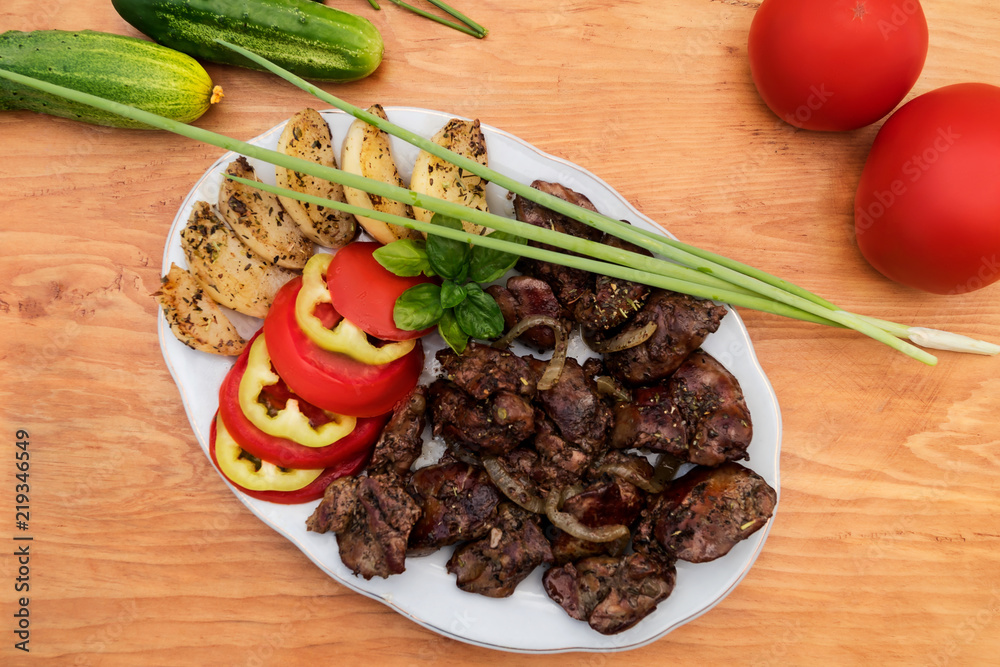 A portion of chicken liver fried in oil along with onions and slices of apples, basil sprig, green onions, tomatoes and cucumbers, all on a wooden background.