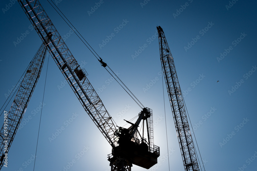 Three construction cranes in silhouette with the sun directly behind and a blue sky, horizontal