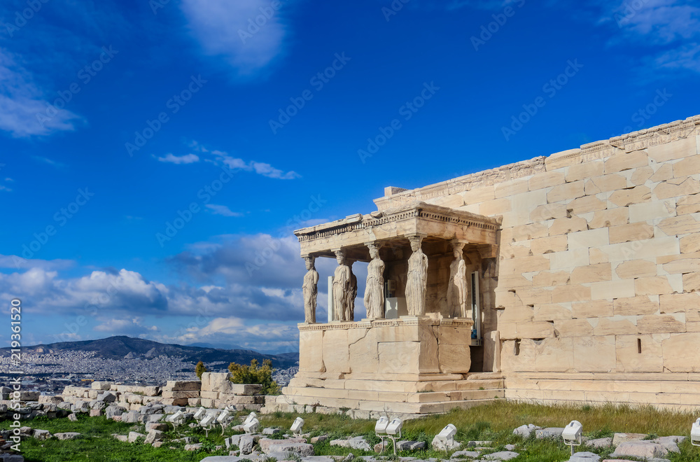 Fototapeta premium View of the Porch of the Caryatids on the Erechtheion temple on the Athens Accropolis with a view of Athens and a mountains in the background under a very blue dramatic cloudy sky