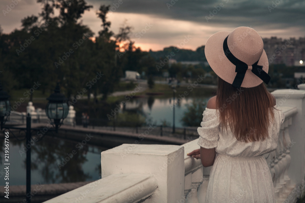 A beautiful young girl in a long white dress and hat walks through the evening park with a pond in the rays of the sunset