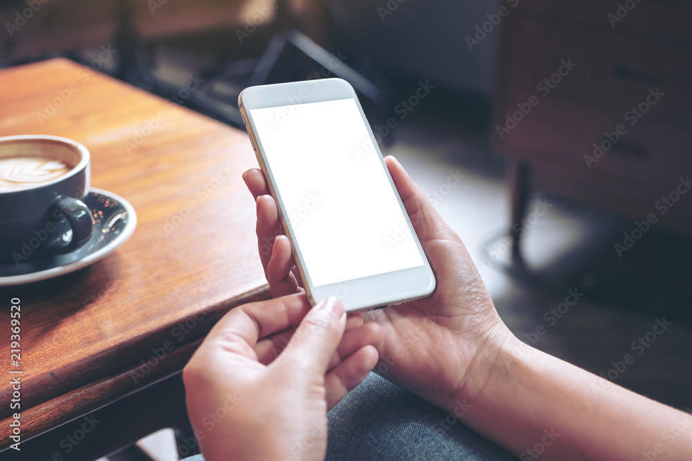 Mockup image of hand holding white mobile phone with blank desktop screen with coffee cup on wooden table in cafe