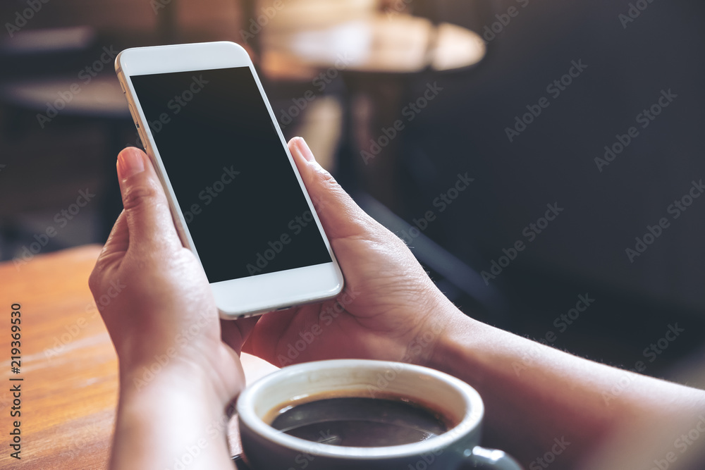 Mockup image of hand holding white mobile phone with blank black desktop screen with coffee cup on wooden table in cafe