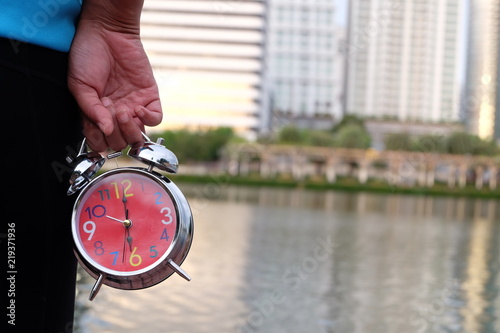 A woman holds a clock in her hand and has a city background.