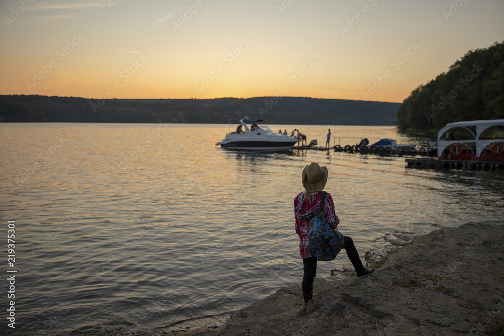 Traveler with a backpack on the back. A girl walks with a backpack. The traveler on the river bank at sunset. Sunset on the river or lake, sea, ocean.