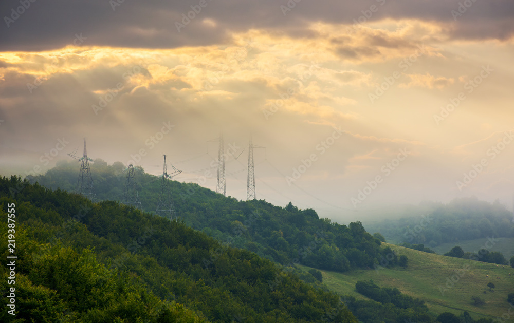 foggy autumn countryside in mountains. power line tower on the forested hill touch the cloud. beautiful golden light