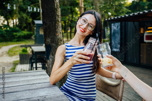 Cheerful brunette woman in eyeglasses and strip dress toasting with her friend outdoor in cafe restaurant in summer park. Holidays, people, party, friendship, fun and vacation concept.