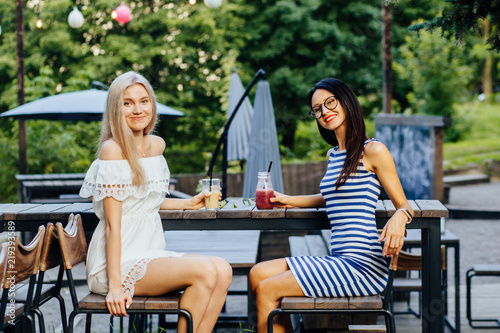 Canvas Print Bright summer portrait of happy sister girls having great time at green outdoor cafe in park, hipster best friends looks on each other, making cheers with tasty cocktails