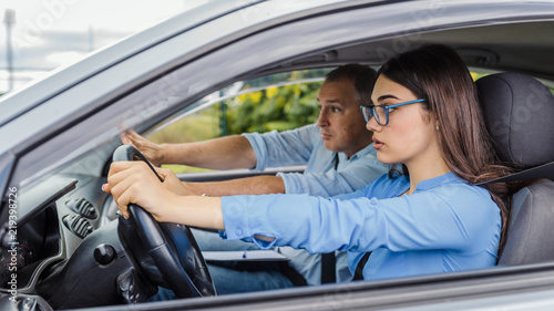 Фотография Young serious inexperienced woman driving a car in alert, worried instructor ma