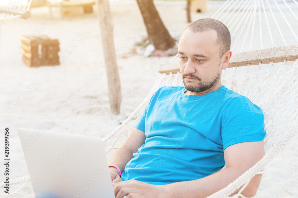 A man with a beard lying in a hammock with a laptop and looks at the screen. Work on the beach while traveling.