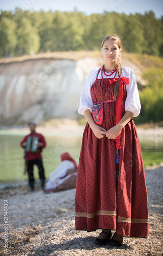 A young beautiful girl in a Russian folk costume stands on the background of a wonderful landscape and her friends