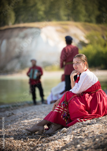 A young girl in a Russian national costume sits against a beautiful landscape and her friends and looks into the distance