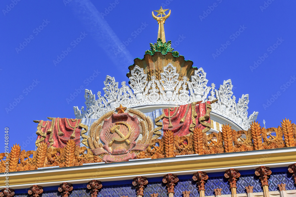 Coat of arms of Ukrainian SSR against gilded spire with soviet star on facade of pavilion on VDNH in Moscow on a blue sky background in sunny summer morning