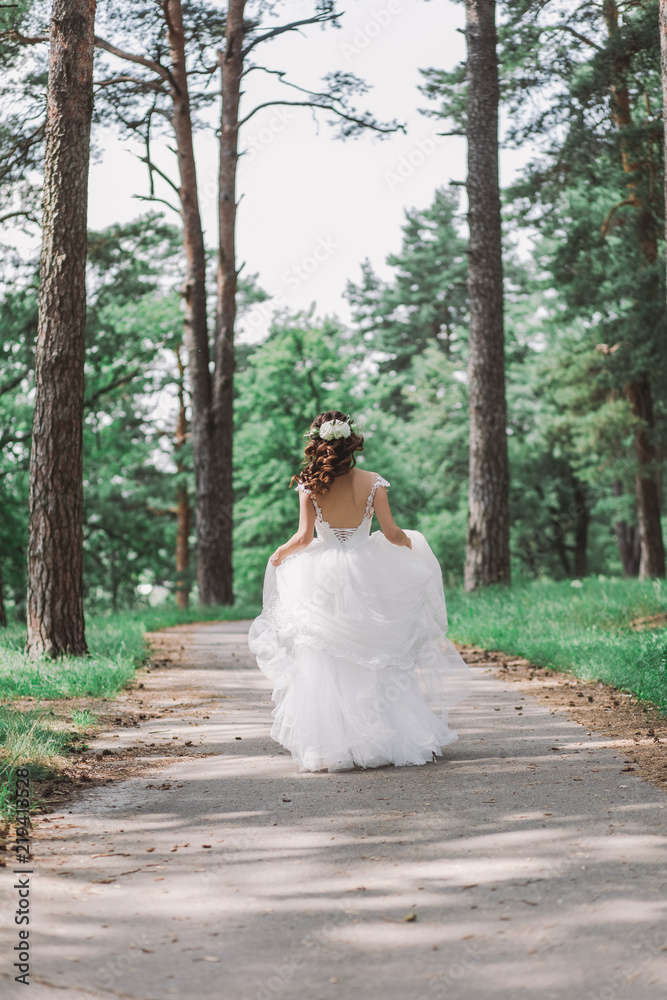 Brunette young bride with long beautiful curly hair. Woman with bridal elegant hairstyle decorated with fresh white flowers walking away from camera outdoors on summer sunny day. Brunette young bride with long beautiful curly hair. Woman with bridal elegant hairstyle decorated with fresh white flowers walking away from camera outdoors on summer sunny day.
