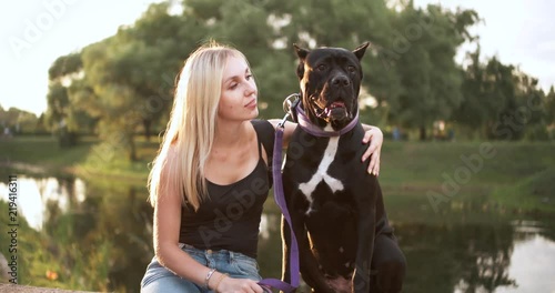Attractive young woman and her dog are sitting on the lake shore in the light of the evening sunset.