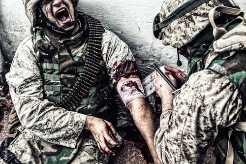 Marine wounded in shoulder, suffering of pain and screaming while receiving medical aid from comrade. Military medic apply pressure bandage to casualty, binding gunshot wound, trying stop bleeding photo