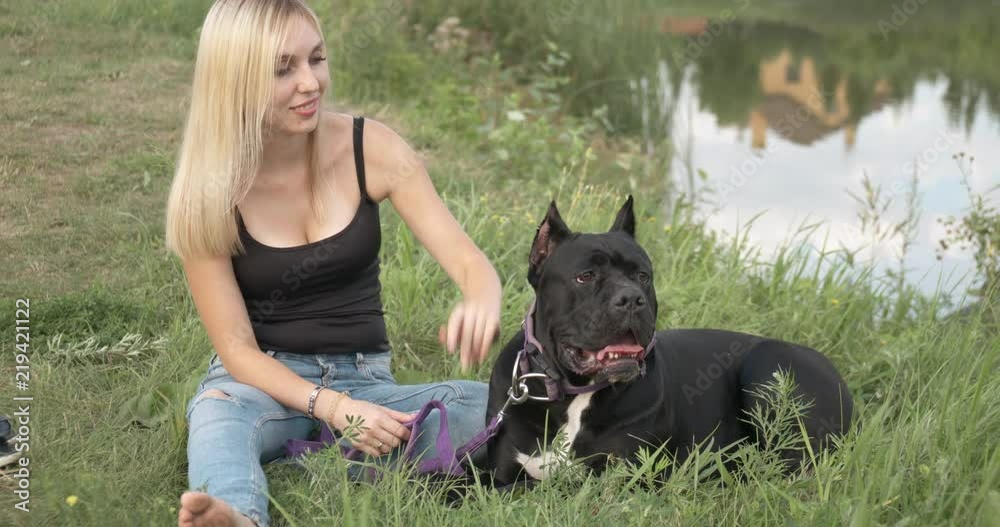 Attractive young woman is sitting on the grass with her dog near the lake. Concept: A dog is a man's best friend, love of animals. Attractive young woman is sitting on the grass with her dog near the lake. Concept: A dog is a man's best friend, love of animals.