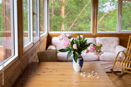 A cozy terrace with furniture with panoramic windows overlooking the forest - a sofa, peonies in a vase on the table and wicker chairs.