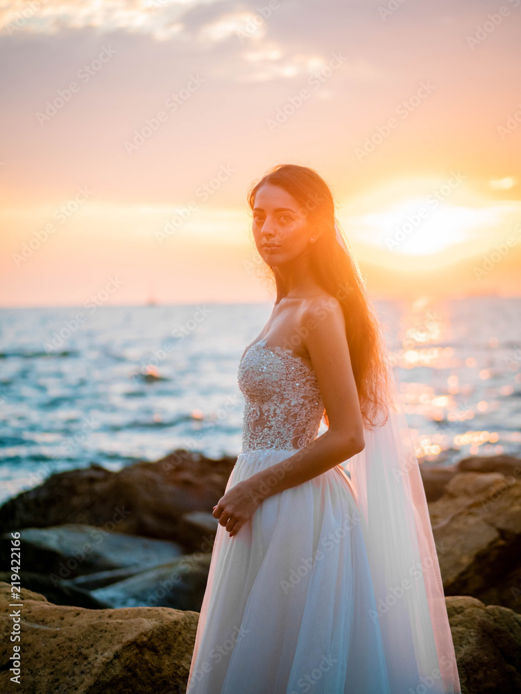 Portrait of girl in wedding luxury dress watching sunrise on sea shore. Bride on a rocks. Beautiful waves near to her. Woman enjoying happy moments with nature. Portrait of girl in wedding luxury dress watching sunrise on sea shore. Bride on a rocks. Beautiful waves near to her. Woman enjoying happy moments with nature.