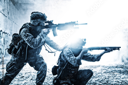 Silhouettes of two army soldiers, U.S. marines team in action, surrounded fire and smoke, shooting with assault rifle and machine gun, attacking enemy with suppressive gunfire during offensive mission photo