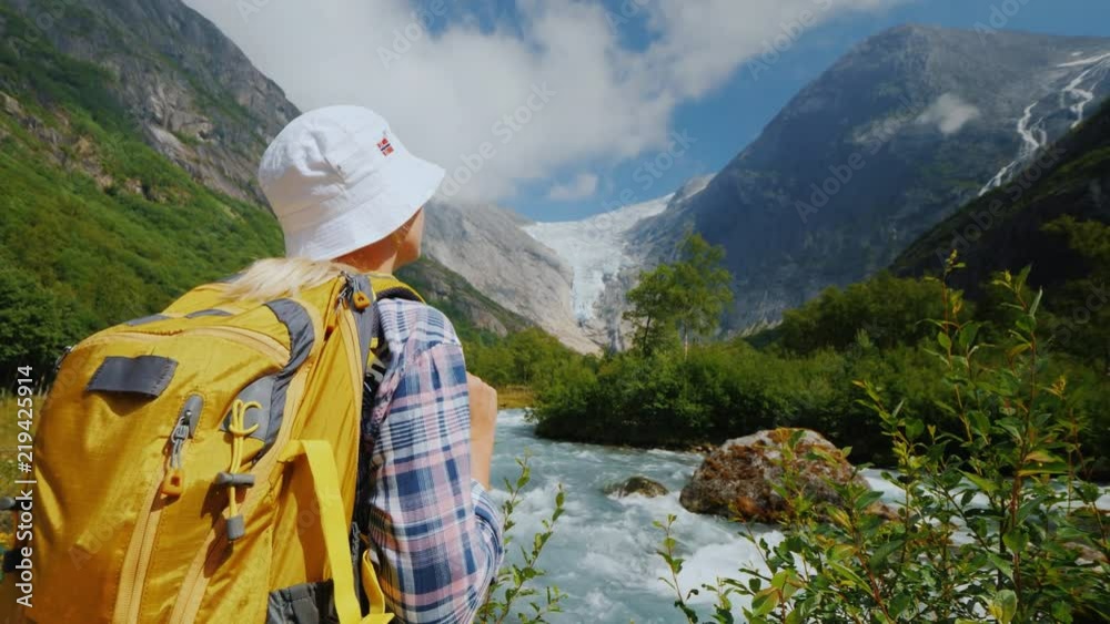 A tourist with a yellow backpack looks at a beautiful glacier at the top of the mountain. Briksdal glacier in Norway A tourist with a yellow backpack looks at a beautiful glacier at the top of the mountain. Briksdal glacier in Norway