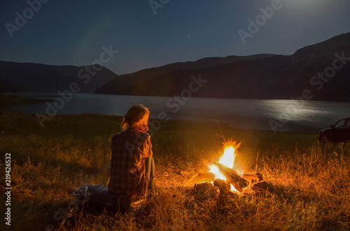 Night camping at sea shore. Female  sitting  at campfire near forest enjoying beautiful view of clear blue water
