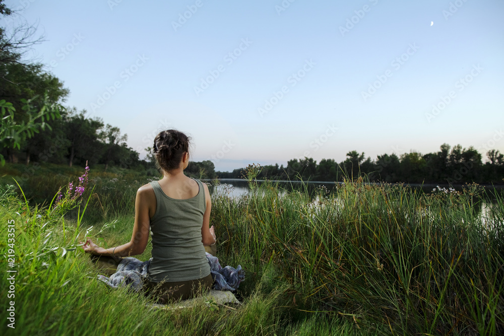 yogin girl sits in a meditative position at sunsat and moon rise on the lake. hands in mudra. Green juicy grass and a calm water surface surrounds it.