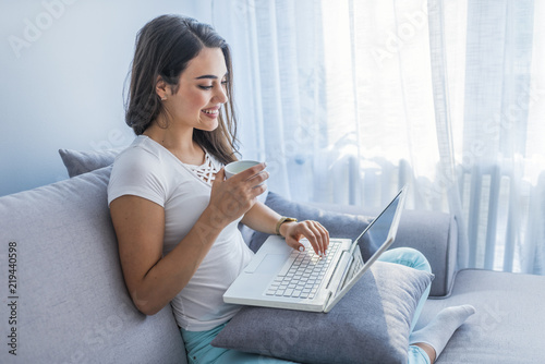 Businesswoman working on laptop computer sitting at home holding a coffee cup in hand. Woman sitting on sofa at home and managing her business.