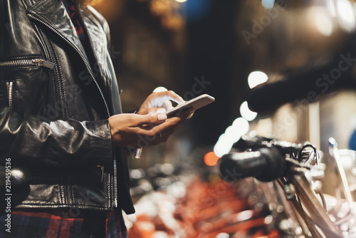 girl in leather jacket using in hands screen smartphone on background bokeh light in night city street, hipster biking and riding to job by ecology  bicycle, lifestyle online wifi internet concept