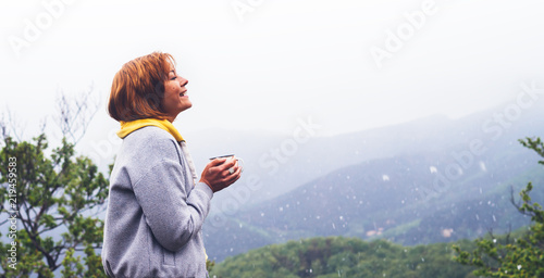 tourist traveler on green top on mountain, girl smiling and holding cup with drink background of falling snow on hills, happy girl enjoying nature panoramic landscape in trip, relax holiday concept