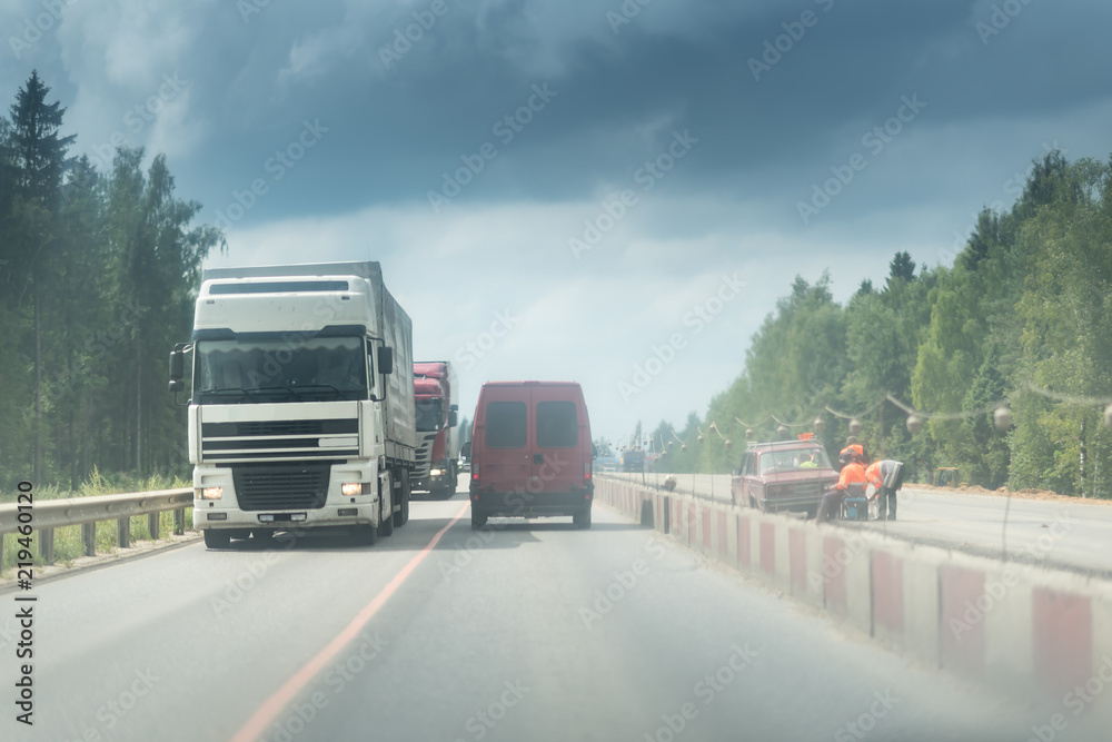 A lorry and truck on the road with concrete white and red blocks from one side and safety rail or barrier from other side. Part of the highway is under construction