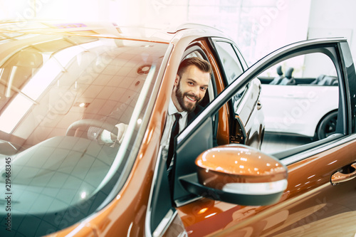 Handsome smiling business man in full suit buying new car in dealership. Salesman shows car photo