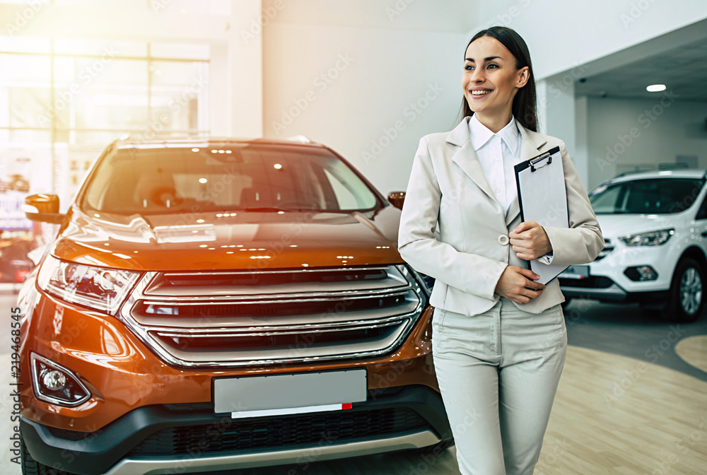 Fototapeta premium Beautiful smiling saleswoman in full suit in dealership on cars background with documents in tablet in hands